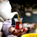 side view of a person pouring black tea from white ceramic teapot into armudu glass